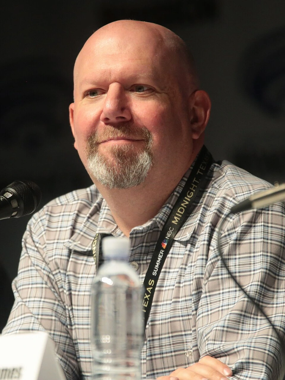 Marc Guggenheim speaking at a panel event, seated behind a microphone and wearing a plaid shirt and lanyard.