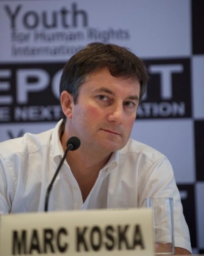 Portrait photo of Marc Koska seated at a conference table with a name placard reading “MARC KOSKA” in front of him.
